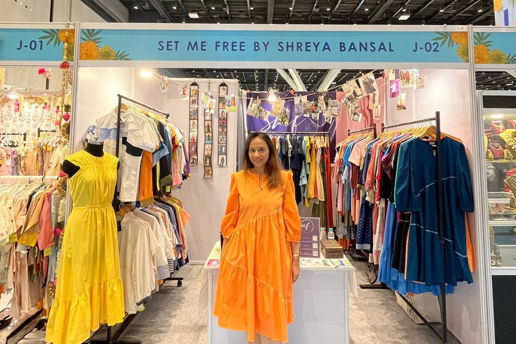 owner in an orange dress standing in a clothing exhibition with various outfits on display.